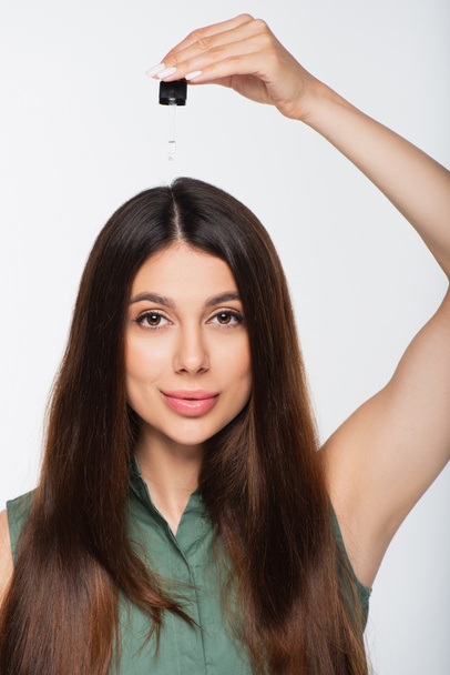jeune femme aux cheveux brillants appliquant de l'huile de traitement sur le dessus de la tête isolé sur gris - Photo, image