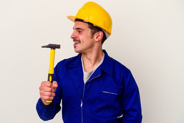 Молодой кавказский рабочий держит молоток, изолированный на белом фоне, смотрит в сторону улыбаясь, веселый и приятный. - Фото, изображение