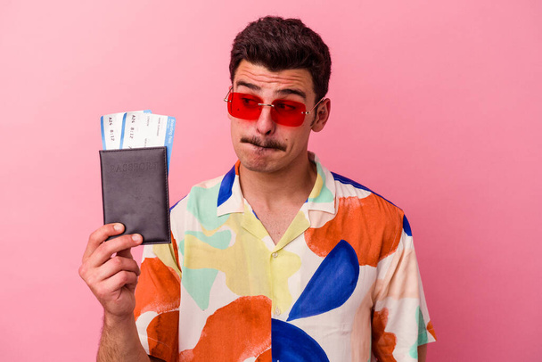 Giovane viaggiatore uomo caucasico in possesso di un passaporto isolato su sfondo rosa confuso, si sente dubbioso e insicuro. - Foto, immagini