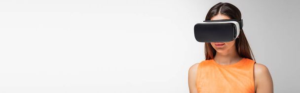 giovane donna bruna in auricolare vr isolato su grigio, banner - Foto, immagini