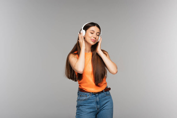 young woman in denim jeans and wireless headphones listening music while smiling isolated on grey - Photo, Image