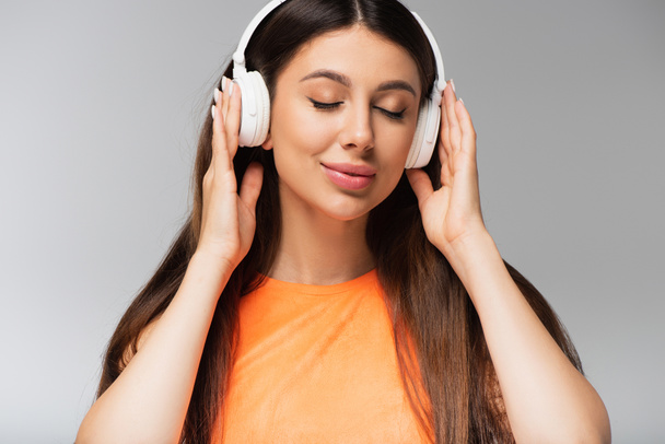 young woman in wireless headphones listening music and smiling isolated on grey - Zdjęcie, obraz