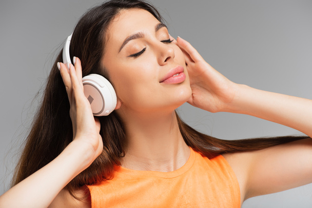ευχαριστημένη νεαρή γυναίκα με ασύρματα ακουστικά που ακούει μουσική απομονωμένη σε γκρι - Φωτογραφία, εικόνα