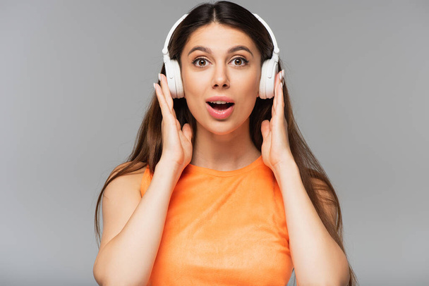 überraschte junge Frau mit drahtlosen Kopfhörern, die isoliert auf grau Musik hört - Foto, Bild
