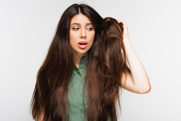 расстроенная молодая женщина, глядя на запутанные и поврежденные длинные волосы, изолированные на сером - Фото, изображение