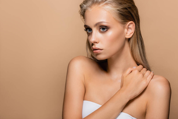 woman with blonde hair and perfect skin posing with hand on naked shoulder isolated on beige - Foto, afbeelding
