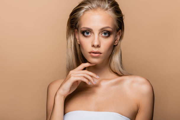 young blonde woman touching chin while looking at camera isolated on beige - Photo, Image