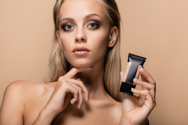woman with blonde hair and bare shoulders holding tube of makeup foundation isolated on beige - Photo, Image