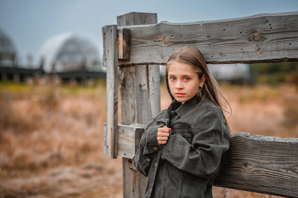 A sad little girl wrapped in an oversized denim jacket standing near a wooden fence. The concept of mental health of adolescents and child loneliness. - Foto, immagini