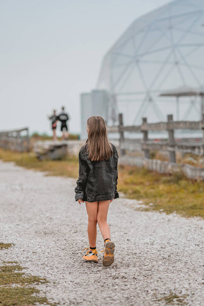 Una niña con una chaqueta de gran tamaño camina sola en un camino en un día nublado - Foto, imagen