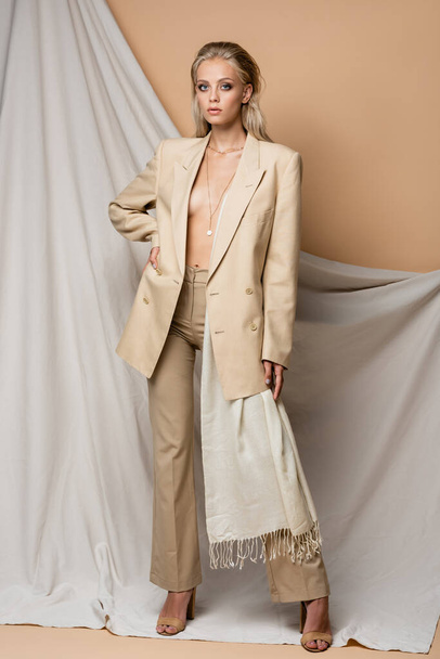 full length view of woman in elegant suit posing with hand on hip near fabric on beige background - Foto, Bild