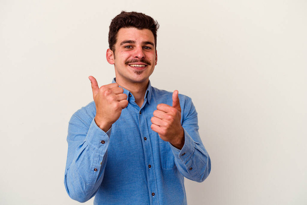 Молодой кавказский человек на белом фоне поднимает оба больших пальца вверх, улыбаясь и уверенно. - Фото, изображение
