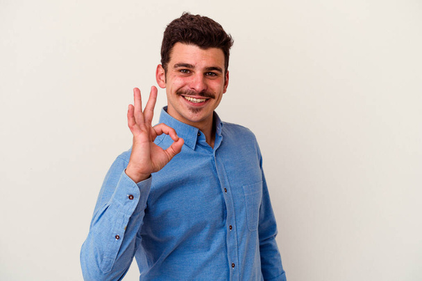 Young caucasian man isolated on white background winks an eye and holds an okay gesture with hand. - Photo, Image