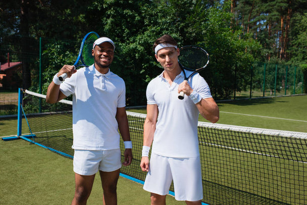Lächelnder afrikanisch-amerikanischer Sportler hält Tennisschläger neben Freund auf dem Platz  - Foto, Bild