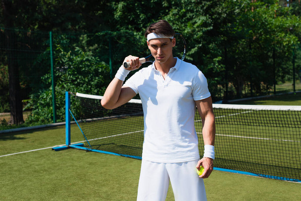 Молодой спортсмен в белой спортивной одежде держит ракетку и мяч на теннисном корте  - Фото, изображение