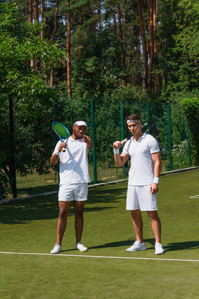 Afroamerikanischer Sportler zeigt mit Finger nahe Freund mit Schläger auf Tennisplatz  - Foto, Bild
