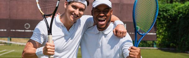 Glückliche interrassische Sportler mit Tennisschlägern, die auf dem Platz in die Kamera schauen, Banner  - Foto, Bild