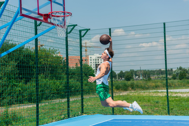 Side view of sportsman jumping under hoop while playing streetball  - Zdjęcie, obraz