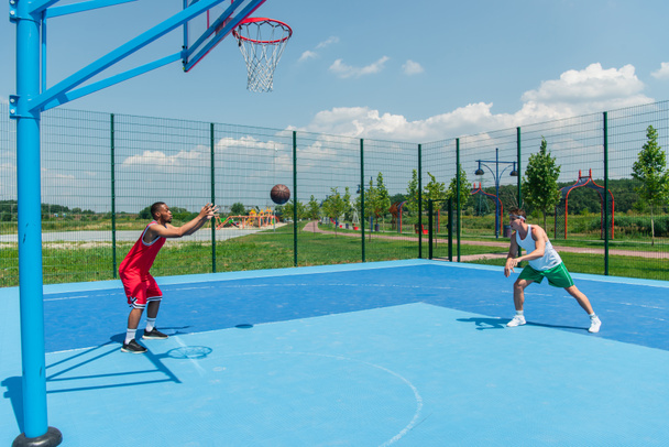 Vista lateral de pelota de baloncesto cerca de deportistas multiétnicos en el patio de recreo  - Foto, Imagen