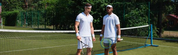 Jugadores de tenis multiétnicos en ropa deportiva blanca con raquetas en la cancha, pancarta  - Foto, imagen