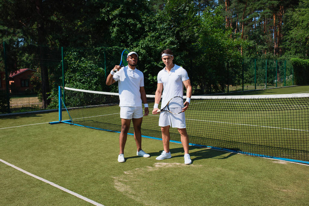 Deportistas multiétnicos sosteniendo raquetas de tenis y mirando a la cámara en la cancha  - Foto, Imagen