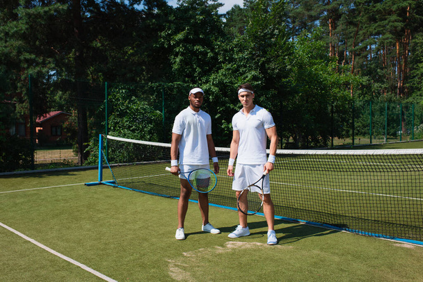 Jóvenes deportistas interracial con raquetas de tenis mirando a la cámara en la cancha  - Foto, Imagen