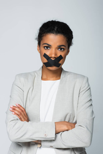 mujer de negocios afroamericana con cinta adhesiva en la boca de pie con los brazos cruzados aislados en gris - Foto, imagen