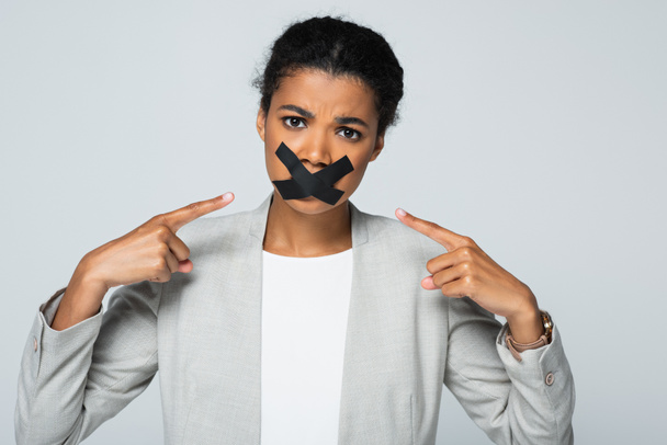 afrikanisch-amerikanische Geschäftsfrau zeigt auf Klebeband auf dem Mund isoliert auf grau - Foto, Bild