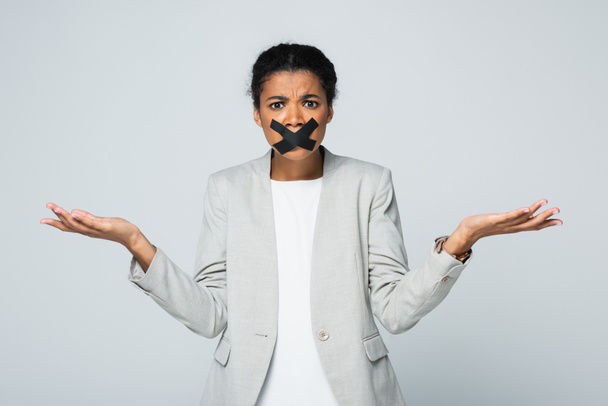 confusa mujer de negocios afroamericana con cinta adhesiva en la boca mostrando un gesto de encogimiento aislado en gris - Foto, Imagen