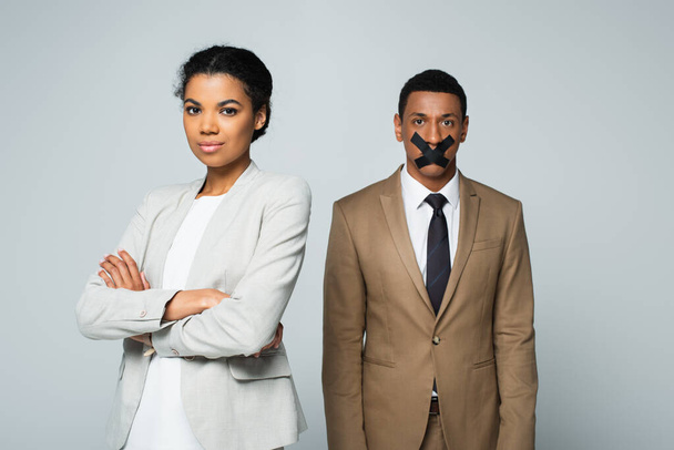 arrogant african american businesswoman standing with crossed arms near manager with scotch tape on mouth isolated on grey - Photo, Image