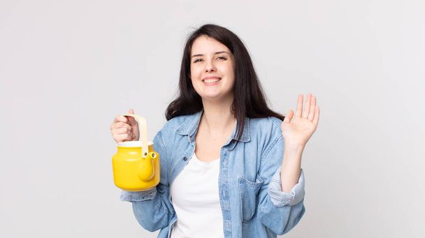 pretty woman smiling happily, waving hand, welcoming and greeting you and holding a teapot - Foto, imagen