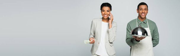 heureuse femme d'affaires afro-américaine tenant des billets en dollars et parlant sur smartphone près du serveur isolé sur gris, bannière - Photo, image