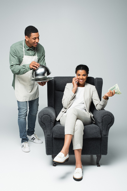 幸せなアフリカ系アメリカ人のビジネスマンドル紙幣を持っていてグレーのウエイターの近くに座っている間スマートフォンで話している女性 - 写真・画像