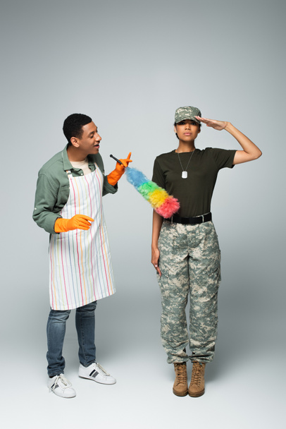 Полная длина женщины-солдата в кепке рядом с любопытным чистильщиком мужского пола в фартуке со шваброй и пыльной щеткой на сером  - Фото, изображение
