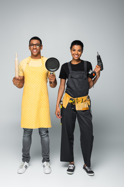 счастливый африканский американец в фартуке держит скалку и сковородку рядом с ремонтницей с электрической дрелью на сером - Фото, изображение