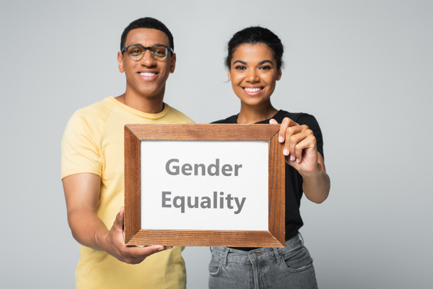 blurred and happy african american man and woman holding wooden frame with gender equality placard isolated on grey  - Zdjęcie, obraz