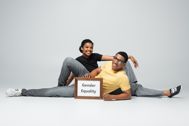 plná délka veselý africký Američan muž a žena sedí v blízkosti plakátu s genderovou rovnost nápisy na šedé - Fotografie, Obrázek