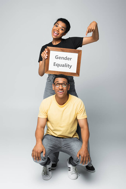 volledige lengte van vrolijke Afrikaans-Amerikaanse vrouw wijzend op bordje met gendergelijkheid belettering in de buurt van man op grijs - Foto, afbeelding