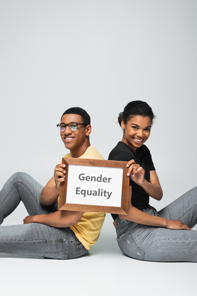 alegre hombre y mujer afroamericanos sentados y sosteniendo pancarta con letras de igualdad de género en gris - Foto, imagen