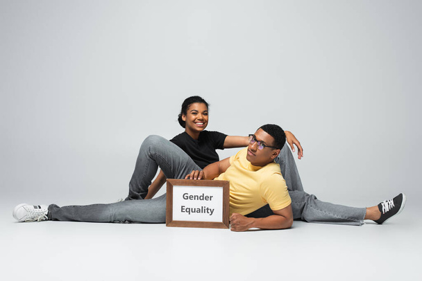 longitud completa de alegre hombre y mujer afroamericanos sentados cerca de pancarta con letras de igualdad de género en gris - Foto, Imagen