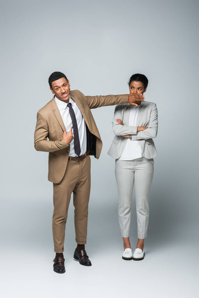 arrogante hombre de negocios afroamericano cubriendo la boca de la mujer de negocios mientras se señala a sí mismo en gris - Foto, Imagen