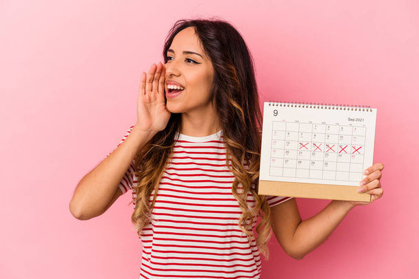 Joven mexicana sosteniendo un calendario aislado sobre fondo rosa gritando y sosteniendo la palma cerca de la boca abierta. - Foto, imagen