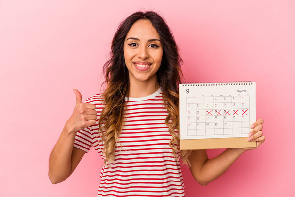 Junge Mexikanerin hält einen Kalender auf rosa Hintergrund und lächelt und hebt den Daumen - Foto, Bild