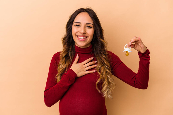 Молодая мексиканская беременная женщина, держащая пустышку, изолированную на бежевом фоне, громко смеется, держа руку на груди.. - Фото, изображение