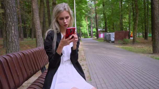 Νέο όμορφο κορίτσι σε ένα λευκό φόρεμα και μια μαύρη ζακέτα χρησιμοποιεί ένα smartphone στη μέση ενός διαδρόμου σε ένα πάρκο ψυχαγωγίας - Πλάνα, βίντεο