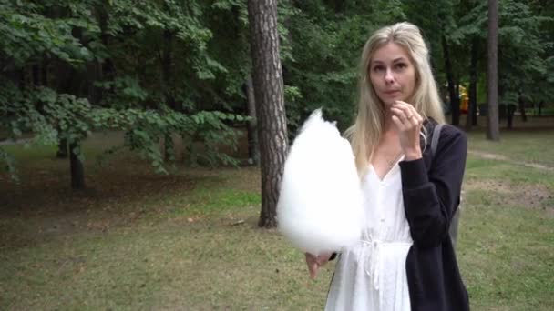 Ein junges schönes Mädchen in weißem Kleid und schwarzer Strickjacke im Park, isst Zuckerwatte und lächelt sehr emotional - Filmmaterial, Video
