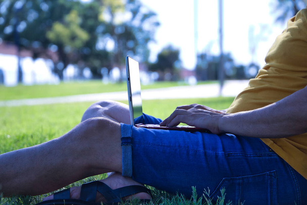 Λεπτομέρεια των ισπανόφωνων ώριμων ενηλίκων χεριών του που κάθονται σε ένα γκαζόν πάρκου, ακουμπώντας σε ένα δέντρο ενώ εργάζεται με το laptop του. - Φωτογραφία, εικόνα