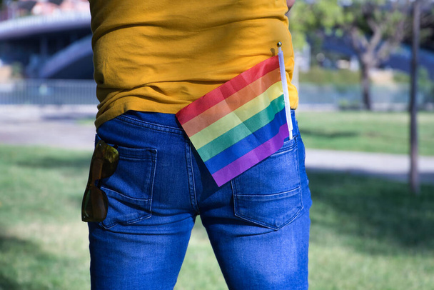 Зрелый взрослый мужчина в футболке горчичного цвета и коротких джинсах с солнцезащитными очками в одном заднем кармане и флагом гей-гордости в другом.. - Фото, изображение