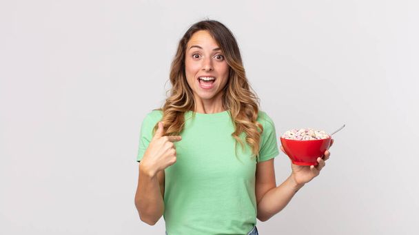 mujer bastante delgada mirando sorprendido y sorprendido con la boca abierta, apuntando a sí mismo y sosteniendo un tazón de desayuno - Foto, imagen