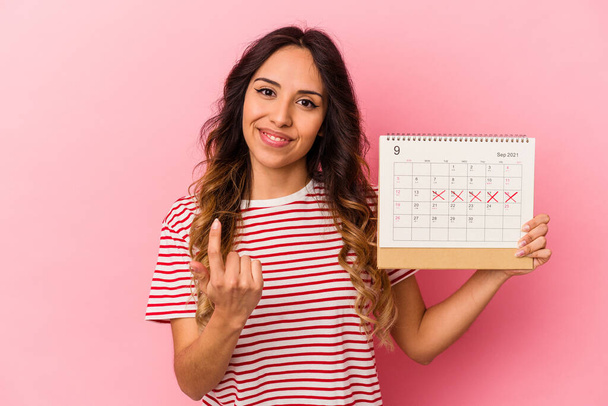 Junge Mexikanerin hält einen Kalender auf rosa Hintergrund und zeigt mit dem Finger auf dich, als ob du einladend näher kommst. - Foto, Bild